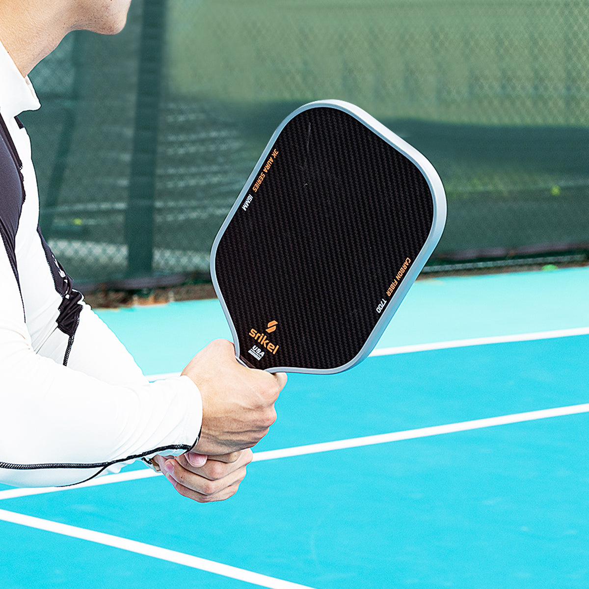 A man wearing a white sleeve holds a Srikel Aura pickleball paddle with a carbon fiber texture on a blue court