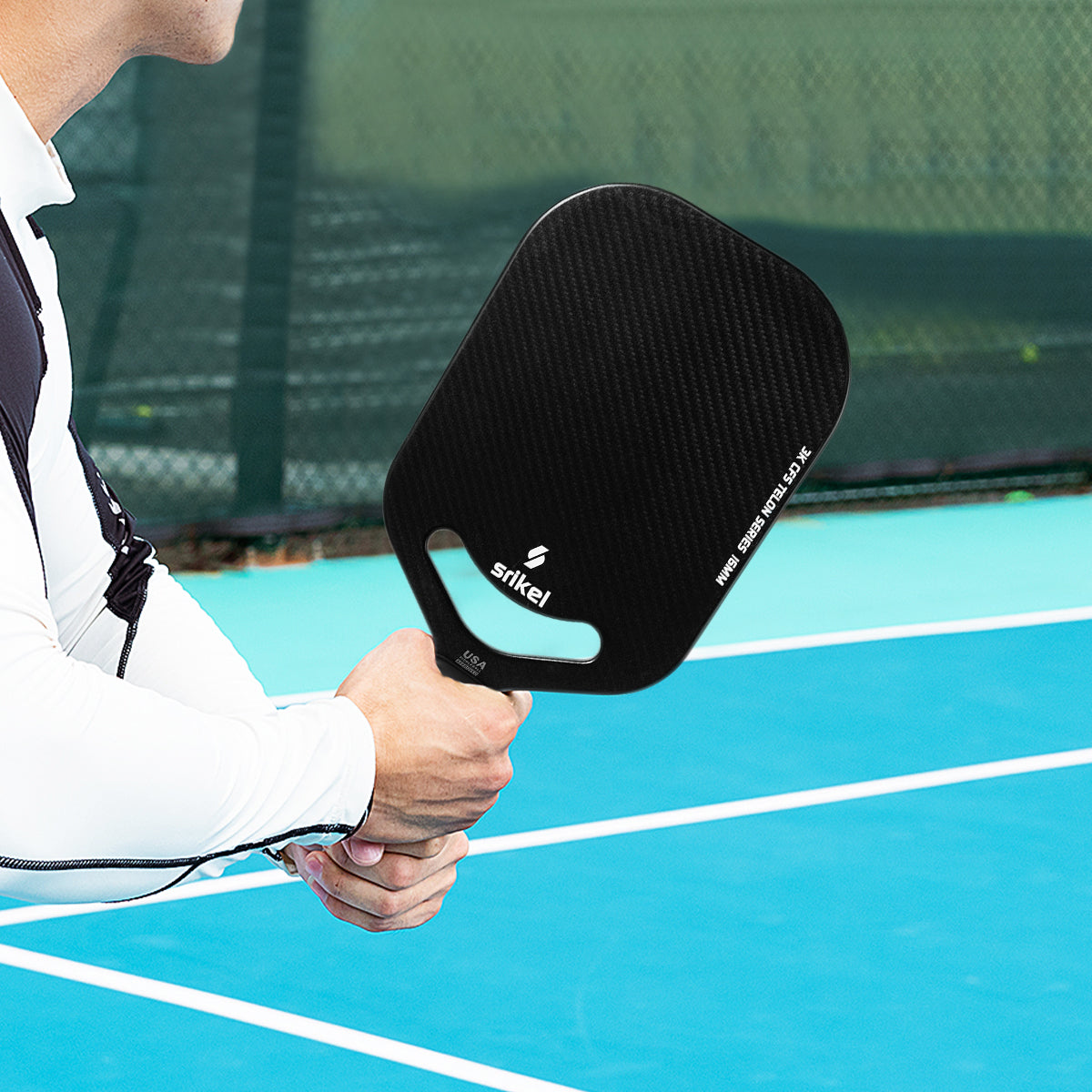 A man wearing a white sleeve plays with a black Srikel Telon pickleball paddle on the court.