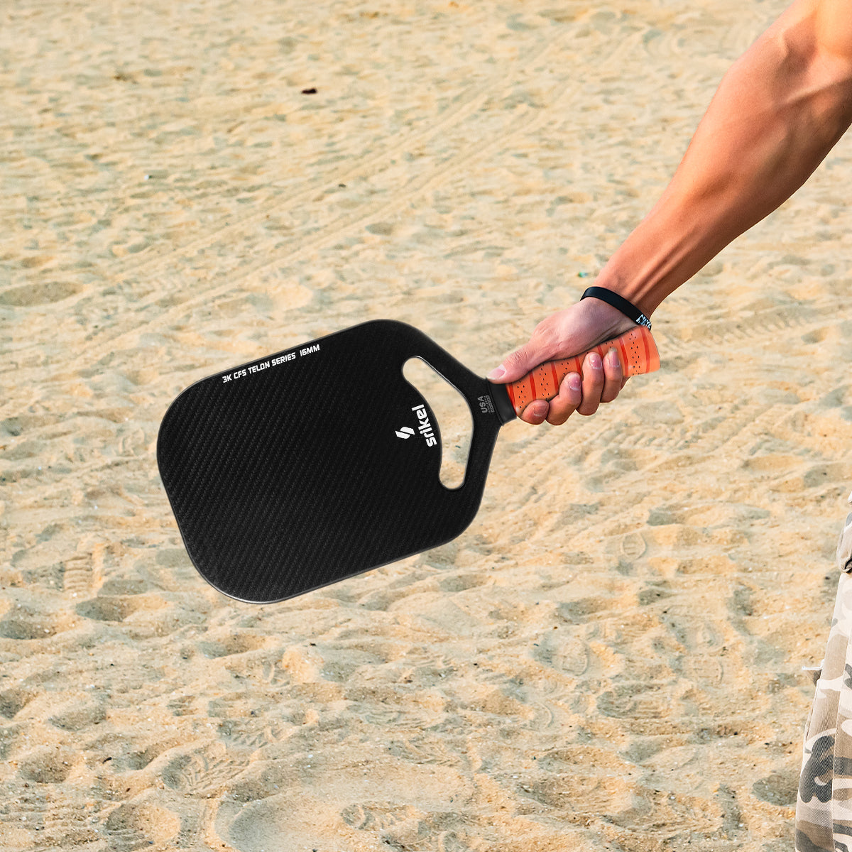 A person holds a Orange color Srikel Telon pickleball paddle with an orange grip on a sandy surface
