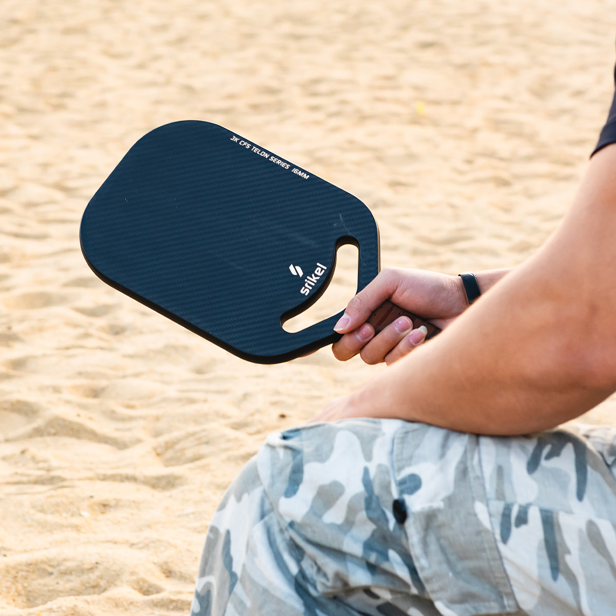 A person sits on sandy ground, holding a black color Srikel Telon pickleball paddle