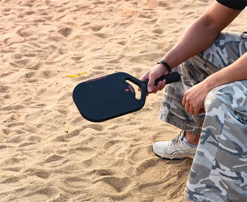 A person sitting on sandy ground holding a black pickleball paddle with a sleek design.