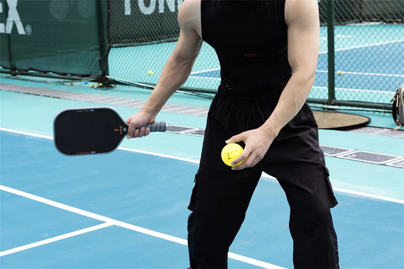 A man in athletic attire holding a paddle and a yellow pickleball, preparing to serve on an outdoor pickleball court.