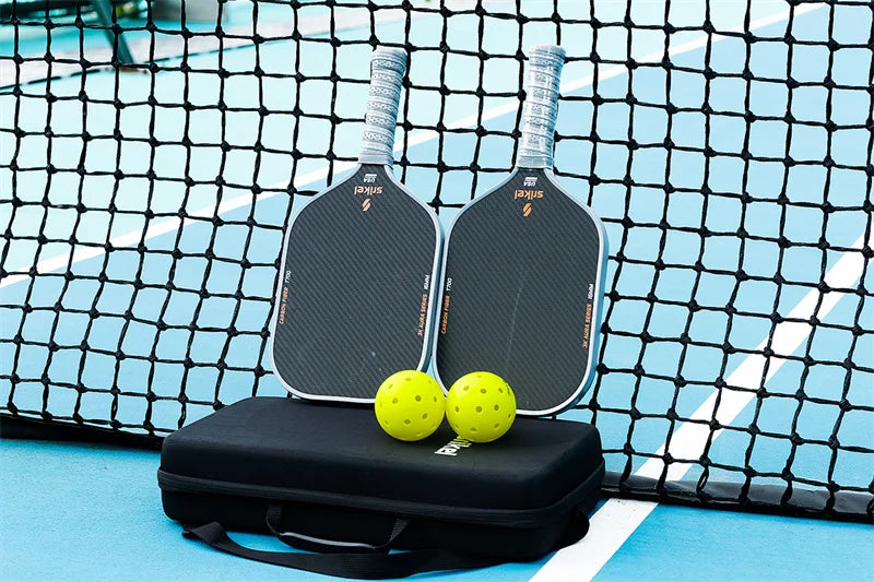 Two pickleball paddles, two yellow pickleballs, and a black carrying case are set against a pickleball court net.