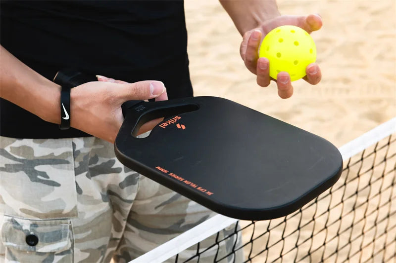 A person holding a Srikel Telon™ pickleball paddle and a bright yellow perforated pickleball near a net on a sandy court.