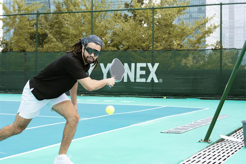 An athlete wearing sunglasses and a headband playing pickleball on an outdoor court.