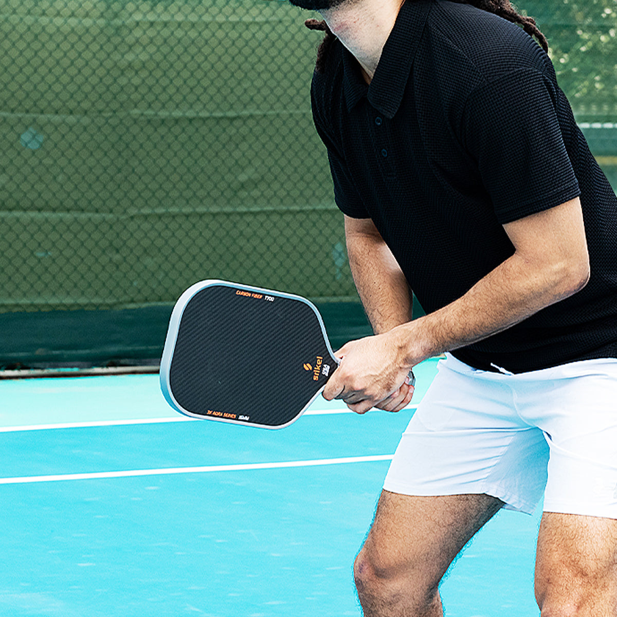 A man in a black shirt and white shorts holds a Srikel pickleball paddle on a blue court