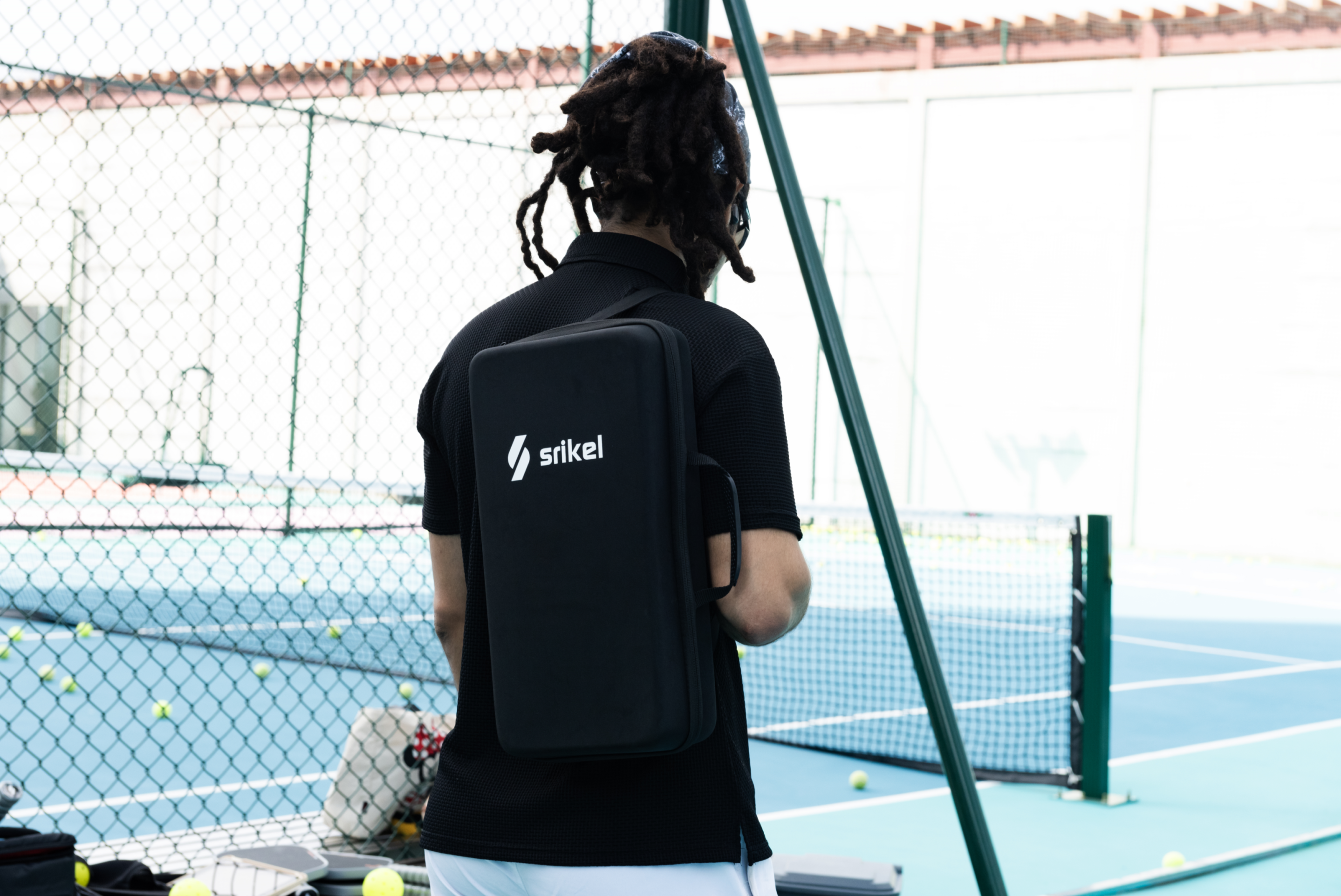 A man is carrying a Strikel Portable Pickleball Sling Bag on their back, standing on a court with pickleball balls scattered around.